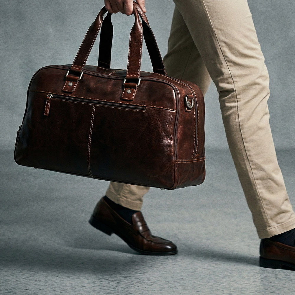 Brown leather duffel bag held by a person wearing beige pants and brown shoes on a gray background