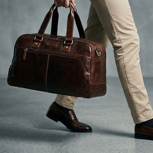 Brown leather duffel bag held by a person wearing beige pants and brown shoes on a gray background