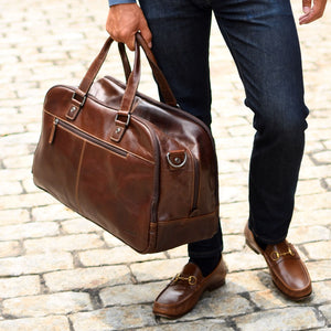 Person holding a brown leather duffel bag on a stone pavement
