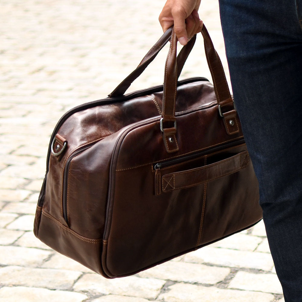 Brown leather duffel bag held by a person on a stone pavement